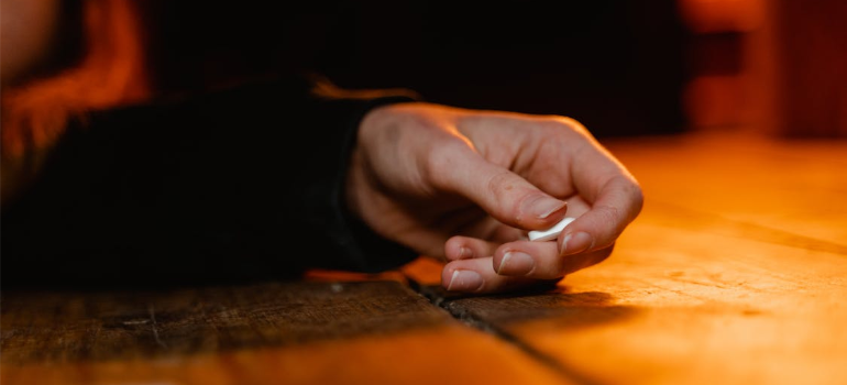 A close-up of a woman’s hand on the floor holding a white pill.