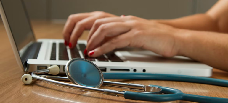 A close-up of a medical professional working on a laptop by a stethoscope.