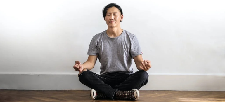 A man in a gray shirt and black jeans meditating on a wooden floor.