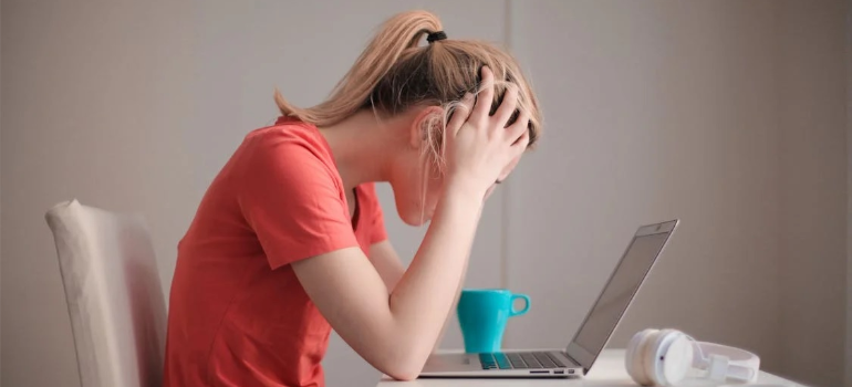A distressed woman holding her head as she works on a laptop.