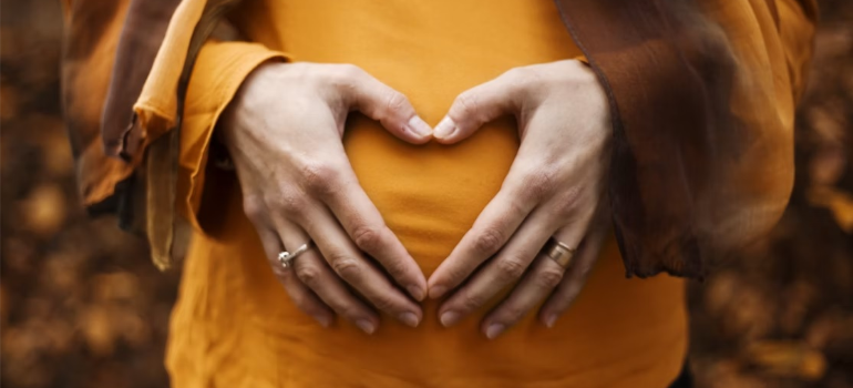 A close-up of a pregnant woman doing a heart shape with her hands over her belly, illustrating how there is no amount of acceptable alcohol during pregnancy.